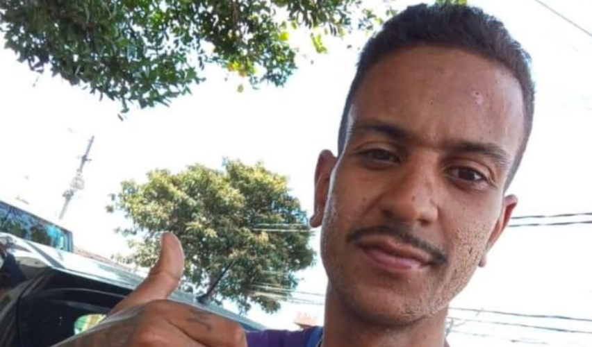 Na imagem, um homem negro de pele parda aparece em primeiro plano, olhando para a câmera e sorrindo levemente. Ele tem cabelo curto, bigode fino e barba rala. Usa camiseta preta e faz um gesto de positivo com a mão direita. Ao fundo, há árvores, fios elétricos e parte de um carro estacionado em uma rua. Justi&ccedil;a condena Estado e ordena novo julgamento de PM que matou sobrinho de rapper em SP