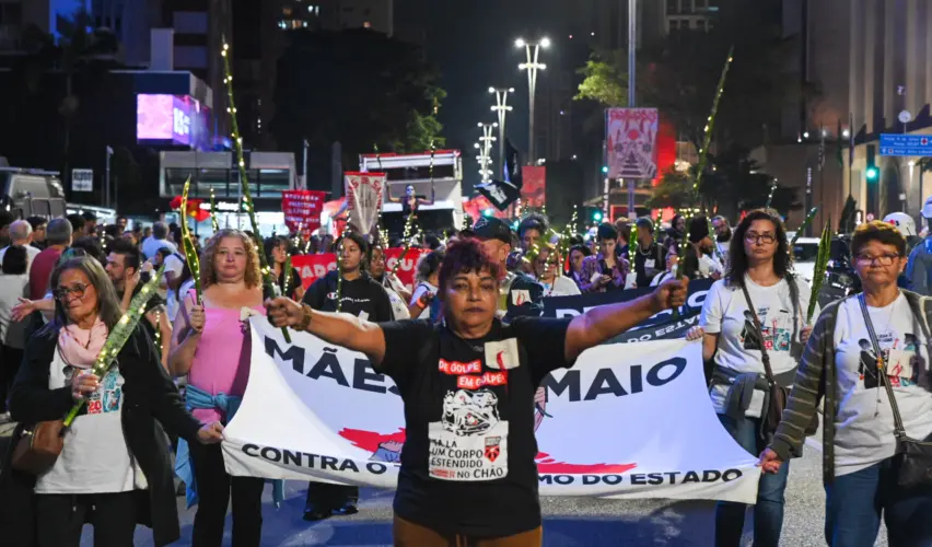 Débora Silva aparece em primeiro plano durante o cortejo do Cordão da Mentira na Avenida Paulista, em São Paulo. Ela caminha segurando uma folha de espada-de-são-jorge em cada mão atrás dela, outras integrantes do movimento seguram uma faixa do Movimento. Débora veste camisa preta está com os braços abertos. “Nossos mortos têm voz”: Cordão da Mentira marca 20 anos dos Crimes de Maio