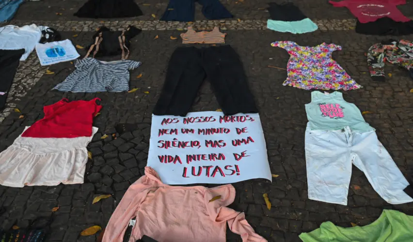 Roupas infantis e adultas estão espalhadas pelo chão em frente ao Parque Trianon Masp, em São Paulo, durante a intervenção “Nossos mortos têm voz”. Entre camisetas, vestidos, uniformes escolares e peças de crianças, um cartaz diz: “Aos nossos mortos, nem um minuto de silêncio, mas uma vida inteira de lutas!”. A instalação homenageia vítimas da violência de Estado representadas pelas roupas deixadas no local. “Nossos mortos têm voz”: Cordão da Mentira marca 20 anos dos Crimes de Maio