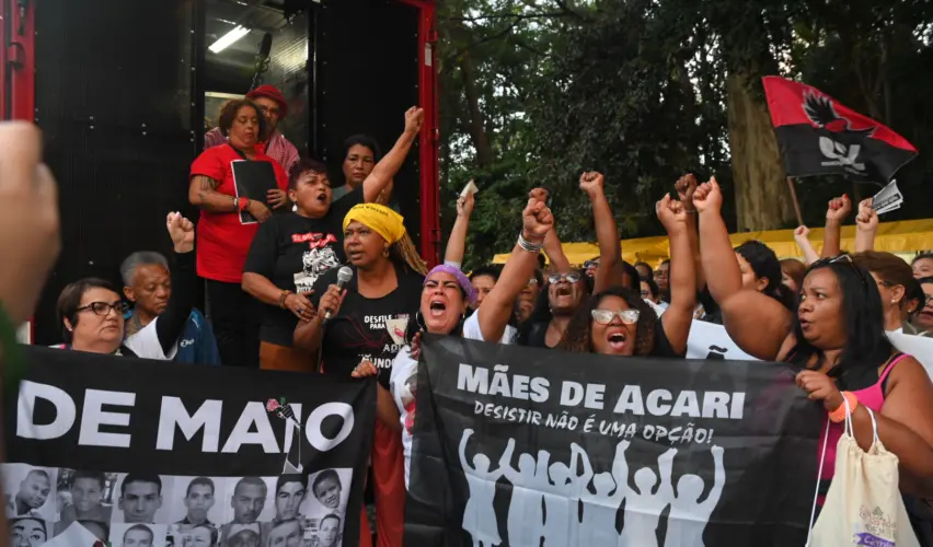 Integrantes de movimentos de mães participam de ato público contra a violência do Estado. Em primeiro plano, mulheres negras erguem os punhos e seguram faixas dos movimentos durante concentração em frente a um caminhão de som “Nossos mortos têm voz”: Cordão da Mentira marca 20 anos dos Crimes de Maio
